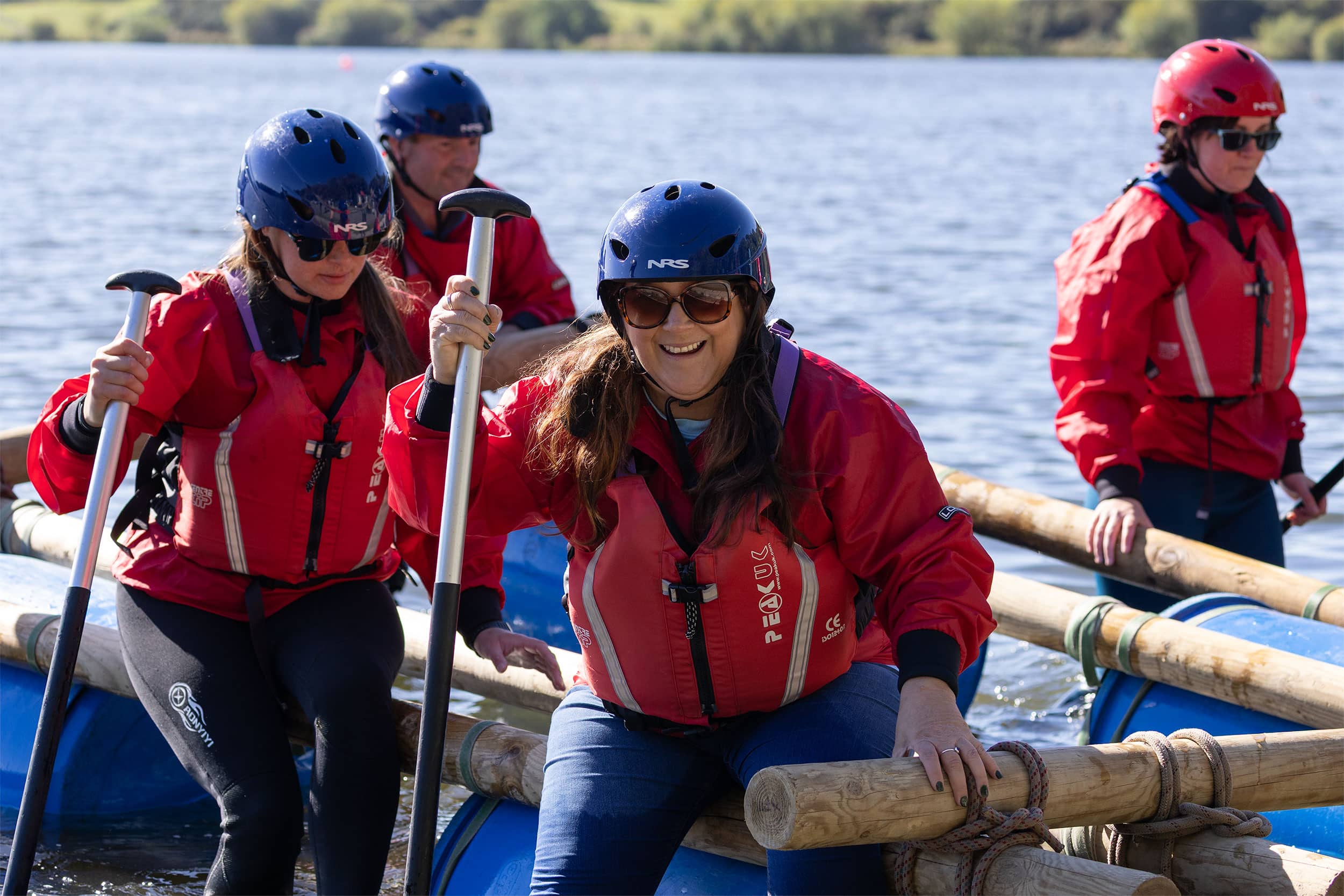 Group of people wearing helmets and life jackets paddling a raft on a lake, captured using commercial photography to showcase outdoor adventure and team-building activities.