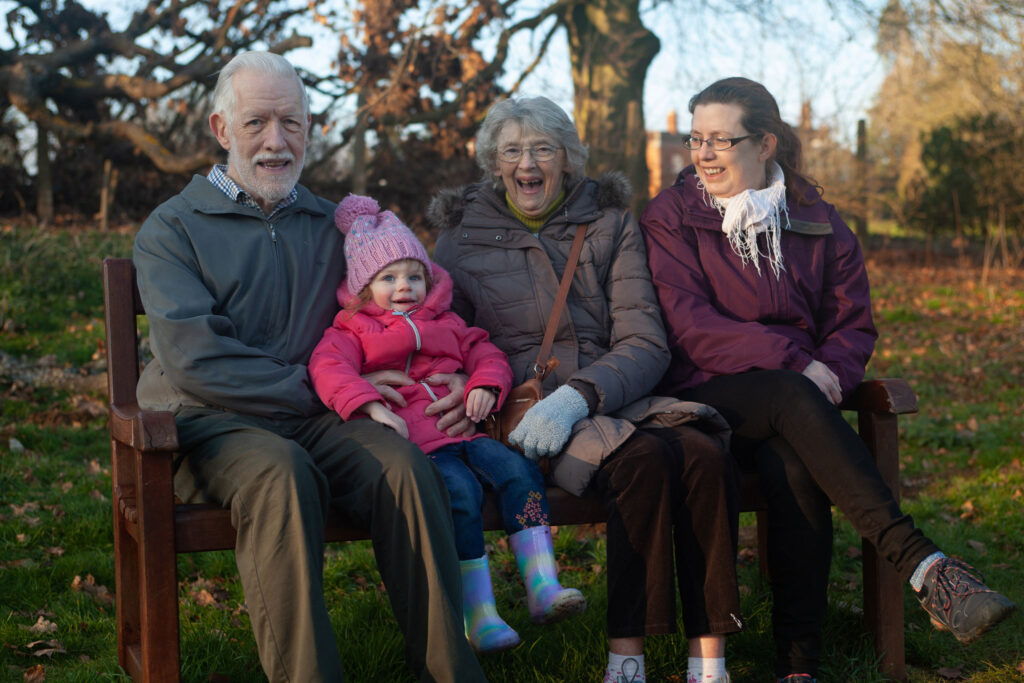 A natural outdoor family portrait, multi-generational family sitting together on a wooden bench outdoors, including grandparents, a young child in a pink coat, and a woman, all smiling in a park setting